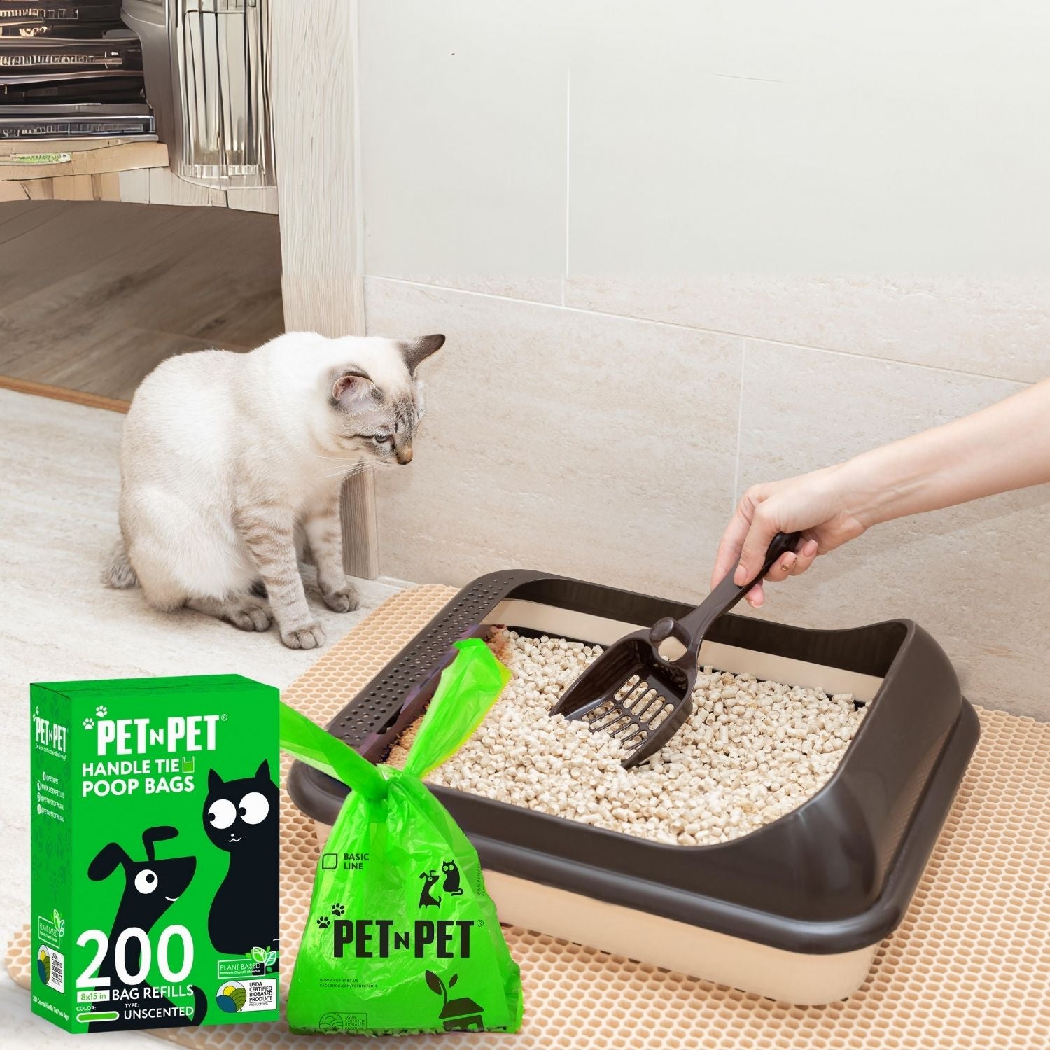 Cat using a litter box with a person cleaning it, next to pet waste bags and a box of pet waste bags.