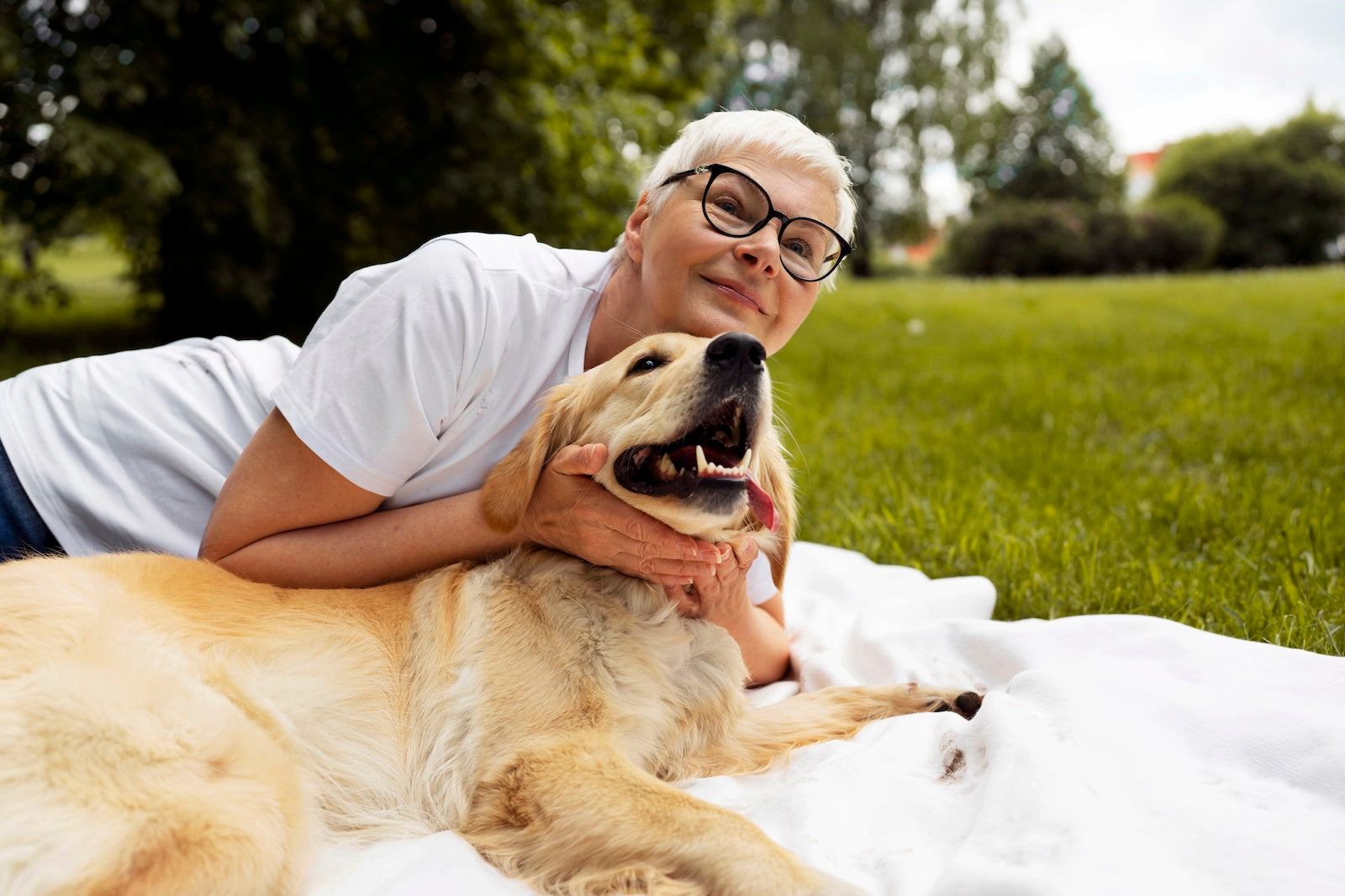 An older woman lying on a blanket outdoors, smiling and hugging her senior golden retriever in a grassy park on a sunny day.